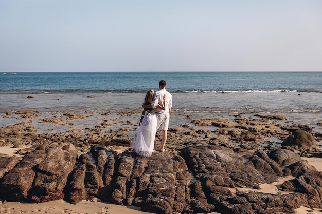 Belo marido e bela esposa se amando em um terreno rochoso e se abraçando, olhando pensativamente para o mar. Conceito de felicidade