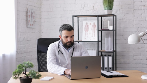 Belo médico trabalhando com laptop em consultório médico