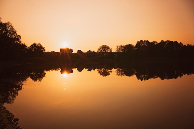 Belo panorama do lago ao pôr do sol Reflexo das casas de sol e árvores em água lisa