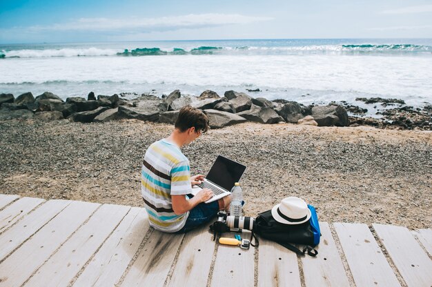 Belo rapaz o fotógrafo a trabalhar com um laptop na praia