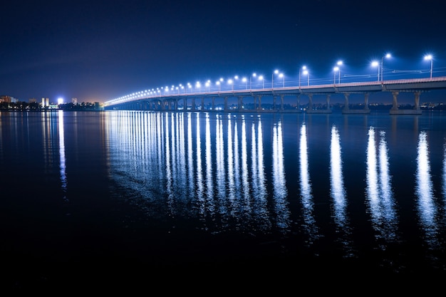 Belo reflexo brilhante de lanternas brilhantes com luz fria no grande rio Dnieper sob uma longa ponte que passa sobre ele