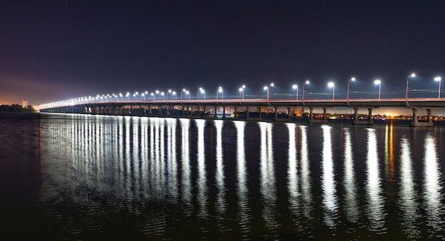 Belo reflexo brilhante de lanternas brilhantes com luz fria no grande rio Dnieper sob uma longa ponte que passa sobre ele