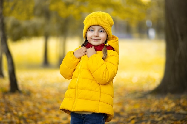 Belo retrato de outono ao ar livre da adorável menina criança de 4 anos, vestindo um casaco e boné amarelo elegante, tocando seu lenço vermelho quente e sorrindo para a câmera.