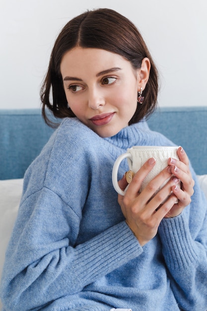 Belo rosto de uma menina, uma menina de camisola azul senta-se em um sofá com uma xícara de chá quente nas mãos dela