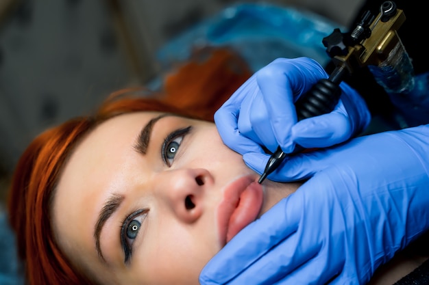 Belo rosto de uma mulher e nas mãos de um cosmetologista fazendo tottoo nos lábios.