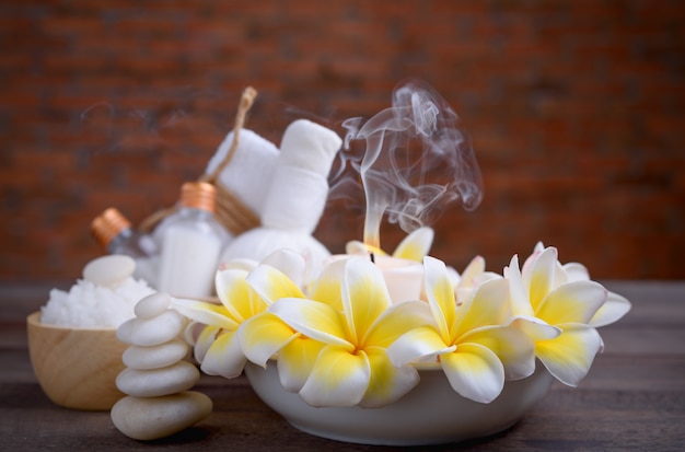 Bem-estar e tratamento de spa com óleos essenciais, pedra zen, toalhas, velas, bola de massagem com flores e flores de frangipani na mesa de madeira com parede de tijolos