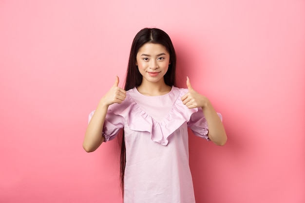 Bem feito. Menina asiática sorridente num vestido mostrando os polegares, gosta e aprova o produto, elogia muito, de pé contra um fundo rosa.