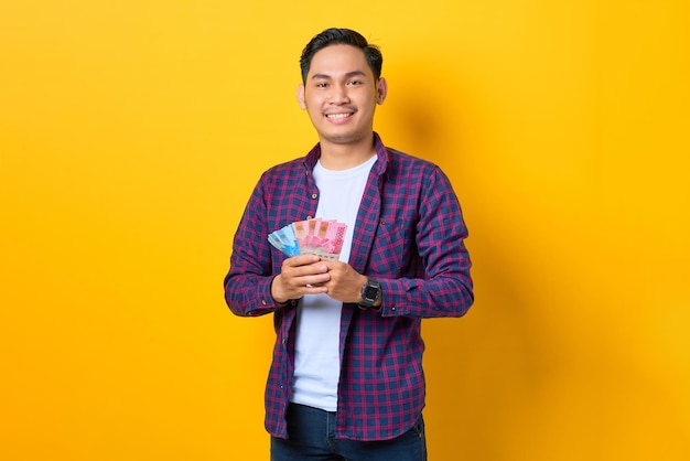 Bem sucedido jovem asiático sorridente em camisa xadrez segurando notas de dinheiro isoladas em fundo amarelo