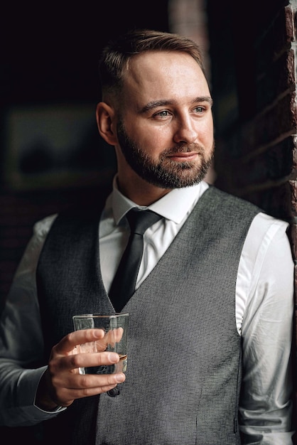 Foto bem sucedido jovem empresário de colete e gravata. com um corte de cabelo estiloso e estiloso e barba. segurando um copo de uísque nas mãos