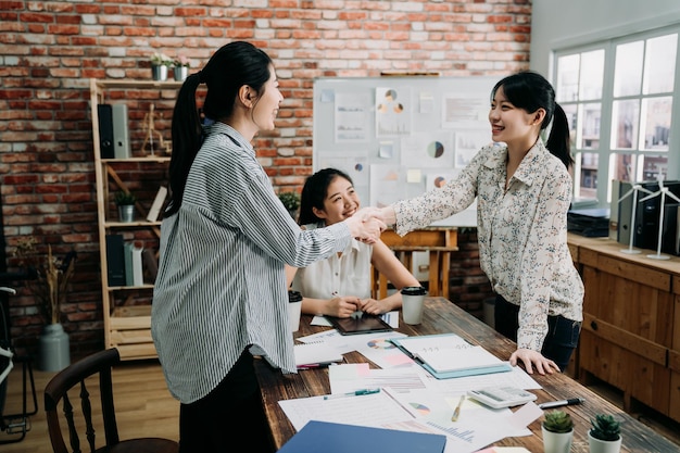 Bem-vindo à nossa equipe. duas jovens mulheres modernas felizes em roupas casuais inteligentes apertando as mãos enquanto trabalhava no escritório criativo na cidade. três trabalhadoras sorridentes lidam juntas com projetos cooperativos.