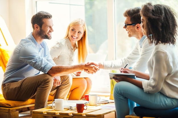 Bem vindo ao nosso time! Duas mulheres jovens felizes trabalhando juntas na área de descanso enquanto dois homens apertam as mãos e sorriem