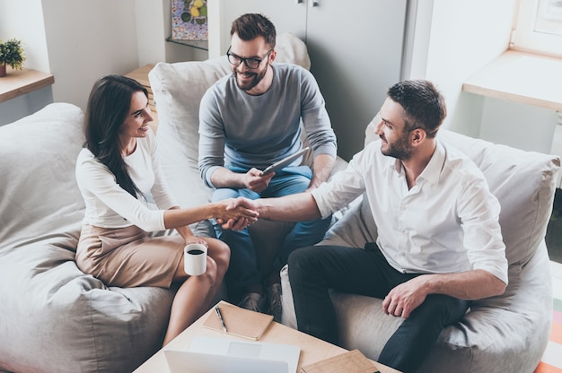 Bem-vindo ao time! Vista superior de três jovens empresários alegres sentados juntos à mesa enquanto um homem e uma mulher apertam as mãos