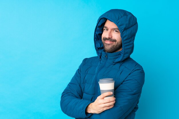 Bemannen Sie die tragende Winterjacke und das Halten eines Mitnehmerkaffees über lokalisierter blauer Wand mit den gekreuzten Armen und vorwärts schauen