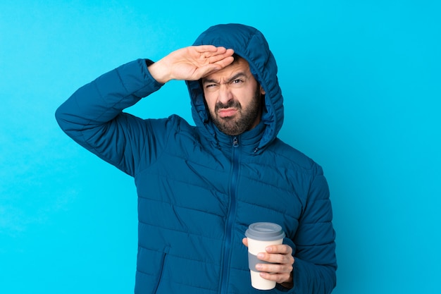 Bemannen Sie die tragende Winterjacke und das Halten eines Mitnehmerkaffees über lokalisierter blauer Wand mit müdem und krankem Ausdruck