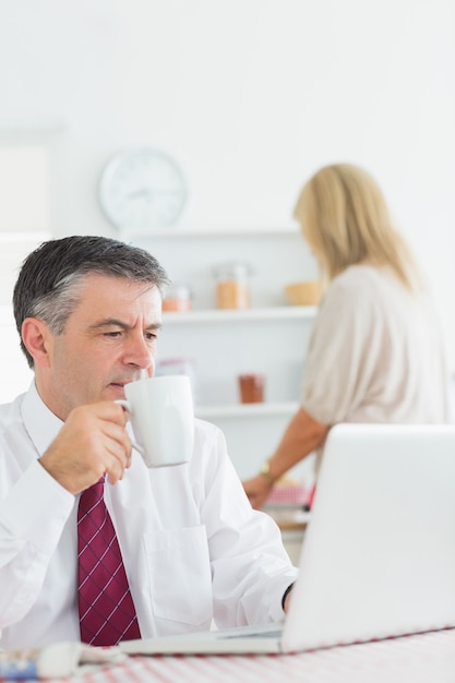 Bemannen Sie trinkenden Kaffee beim Überprüfen des Laptops in der Küche vor der Arbeit
