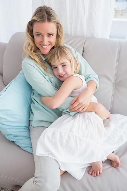 Bemuttern Sie das Sitzen auf der Couch mit ihrer Tochter, die an der Kamera lächelt