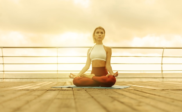 Üben Sie morgens Yoga im Freien. Junge Frau Yogi in Sportkleidung sitzt bei Sonnenaufgang im Lotussitz am Strand. Innere Harmonie