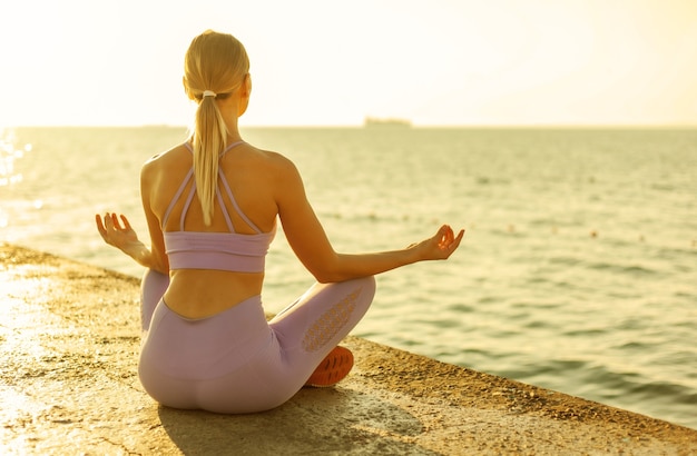 Üben von Yoga. Meditation bei Sonnenaufgang. Junge schlanke Frau sitzt im Lotussitz am Strand. Gesundes Lebensstilkonzept