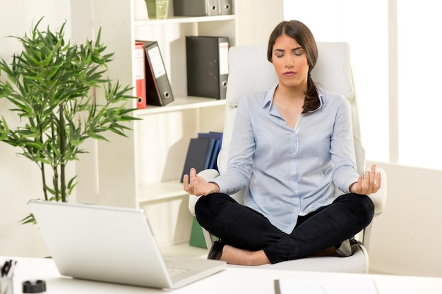 Übendes Yoga der schönen jungen Geschäftsfrau im Büro. Sie sitzt im Lotussitz auf einem Stuhl vor ihrem Schreibtisch.