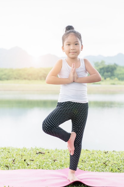 Übendes Yoga des kleinen netten asiatischen Mädchens werfen auf einer Matte im Park-, gesundem und Übungskonzept auf