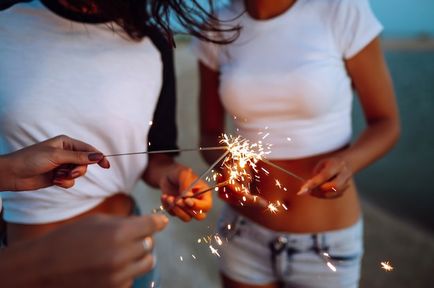 Las bengalas en las manos de chicas jóvenes en la playa. Tres chicas disfrutando de fiesta en la playa con bengalas. Concepto de vacaciones de verano, vacaciones, relax y estilo de vida.