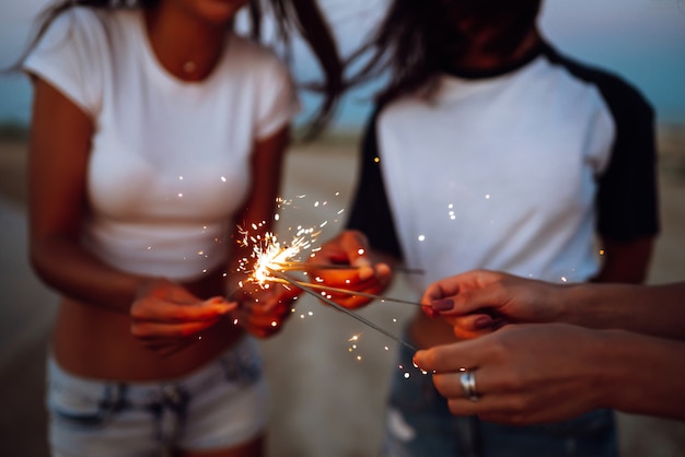 Las bengalas en las manos de chicas jóvenes en la playa. Tres chicas disfrutando de fiesta en la playa con bengalas. Concepto de vacaciones de verano, vacaciones, relax y estilo de vida.