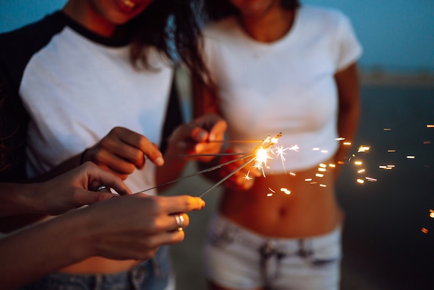 Las bengalas en las manos de chicas jóvenes en la playa. Tres chicas disfrutando de fiesta en la playa con bengalas. Concepto de vacaciones de verano, vacaciones, relax y estilo de vida.