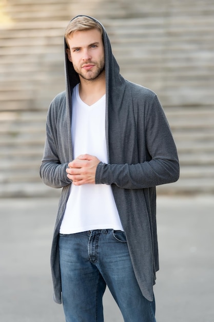 Bequeme Kleidung für den täglichen Gebrauch Toller Geschmack, um sich gut zu kleiden Männlicher Modeeinfluss Modischer junger Modellmann Streetstyle-Outfit Schöner Mann mit Kapuze im städtischen Hintergrund Modetrend