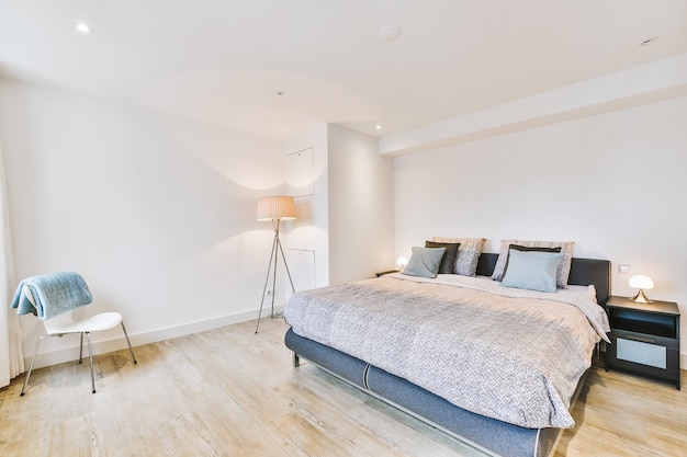 Bequemes Bett mit Decke und Kissen in der Nähe von Lampen und Stuhl im geräumigen Schlafzimmer in moderner Wohnung
