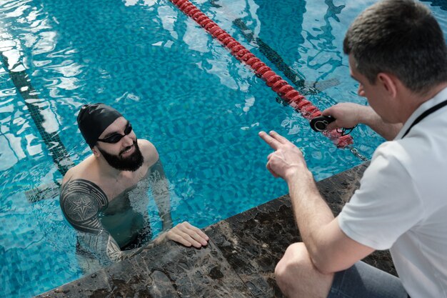 Über Schulteransicht des Trainers mit Stoppuhr gestikulierender Hand, während Schwimmer im Pool lobend