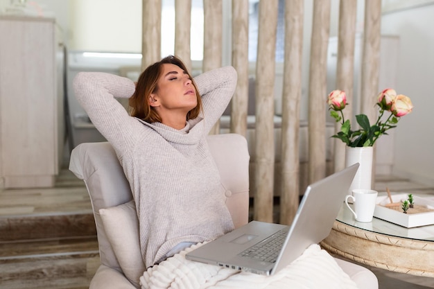 Überarbeitete junge Frau, die in ihrem Haus mit einem Laptop vor ihr sitzt und ihre Arme mit geschlossenen Augen über den Kopf streckt, um die Muskeln zu entspannen, Spannungen abzubauen, das Sehvermögen zu verbessern,