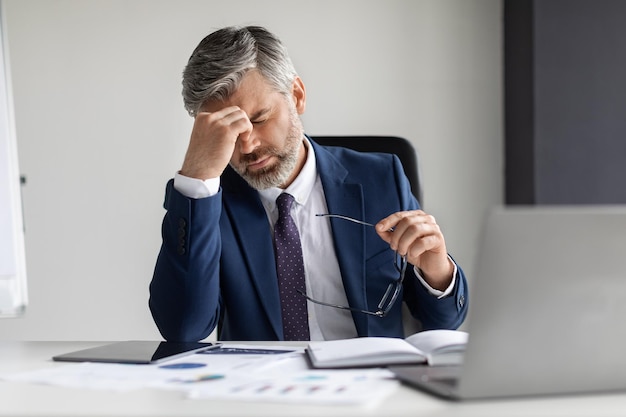 Überarbeitungskonzept Müder reifer Geschäftsmann, der während der Arbeit im Büro unter Kopfschmerzen leidet