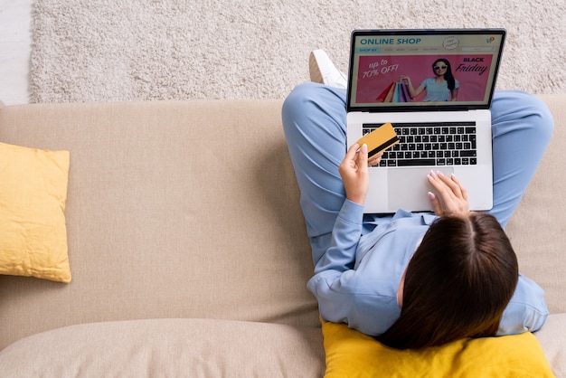 Überblick über junge brünette Frauen im blauen Pyjama, die auf der Couch im Wohnzimmer sitzen und mit Karte für Waren aus dem Online-Shop bezahlen