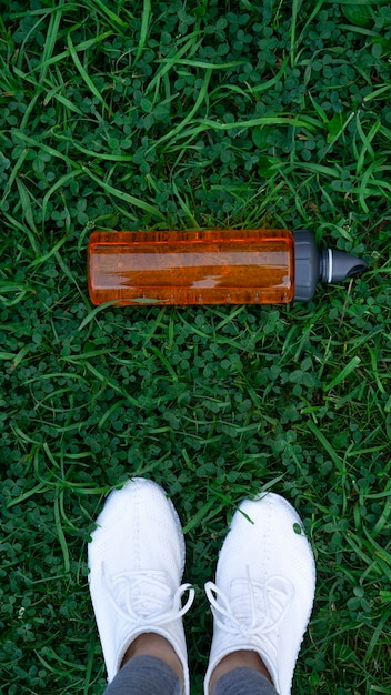 Bereit für das Training. Draufsicht auf einen Grashintergrund mit einer orangefarbenen Sportwasserflasche und weißen Turnschuhen. Langlaufen, Joggen, Bewegung an der frischen Luft.
