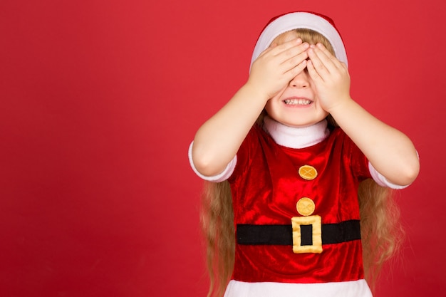 Bereit für Geschenke. Halbe Länge Aufnahme eines kleinen hübschen Mädchens im Weihnachtsmann-Outfit, das breit lächelt und die Augen mit den Händen schließt