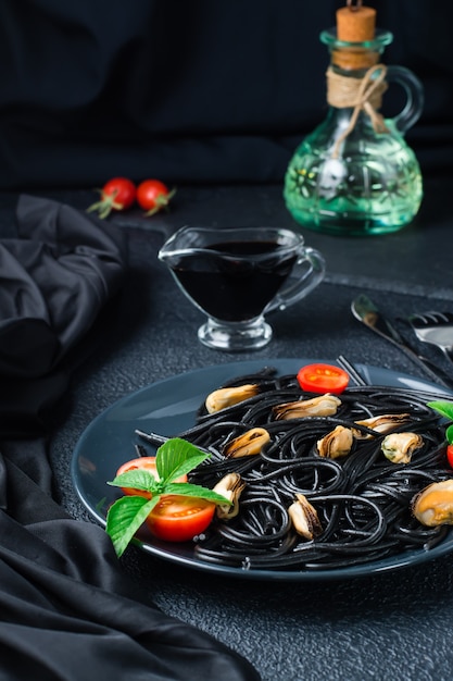 Bereit, schwarze Spaghetti mit Muscheln, Tomaten und Basilikum auf einem Teller und Sojasauce auf schwarzem Hintergrund zu essen. Foodfotografie in dunklen Farben. Vertikale Ansicht