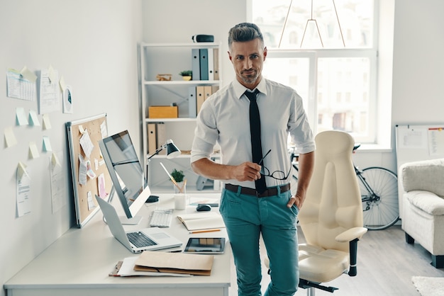 Bereit zu arbeiten. Gut aussehender junger Mann, der in die Kamera schaut und lächelt, während er im Büro steht