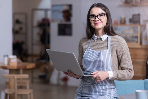 Bereit zu arbeiten. Schöne ehrgeizige Frau, die lächelt und Laptop hält, während sie in einem Café steht.