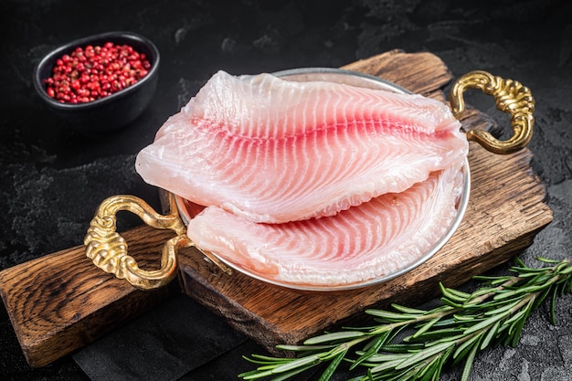 Bereit zum Kochen von rohem Tilapia-Fischfilet in einer Pfanne mit Kräutern Schwarzer Hintergrund Draufsicht