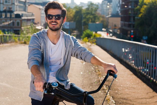 Bereit zum reiten. Schöner junger Mann auf dem Fahrrad, der in die Kamera schaut und lächelt, während er im Freien steht