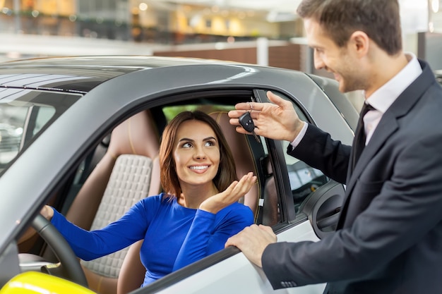 Bereit zur Probefahrt. Hübscher junger Autoverkäufer, der der jungen schönen Frau den Schlüssel gibt