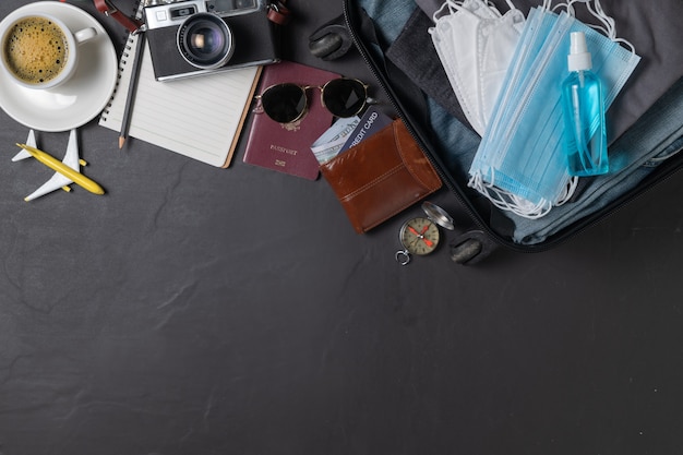 Bereiten Sie Maske und Alkoholspray in Koffer, Vintage-Kamera, Notizbuch, Karte auf schwarzem Tisch und Kopierraum vor. Reisen und Covid-19-Konzept verhindern