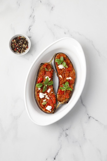 Berenjenas al horno rellenas con salsa de tomates picados en un plato al horno y fondo de mármol blanco