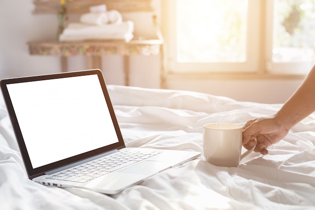 Übergeben Sie das Halten der Kaffeetasse und des Laptops auf dem Bett in der Morgenzeit, Fokus an Hand