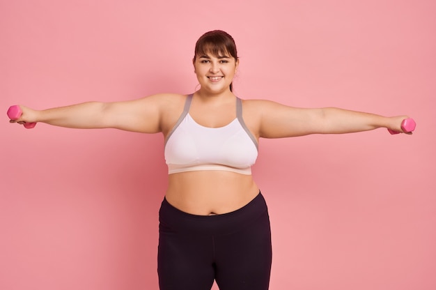 Übergewichtige Frau, die Übung mit Hanteln, rosa Wand, Körper positiv tut