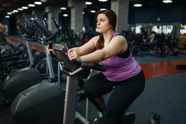 Übergewichtige Frau trainiert auf Heimtrainer im Fitnessstudio, aktives Training. Übergewichtige weibliche Person kämpft mit Übergewicht, Aerobic-Training gegen Fettleibigkeit, Sportverein