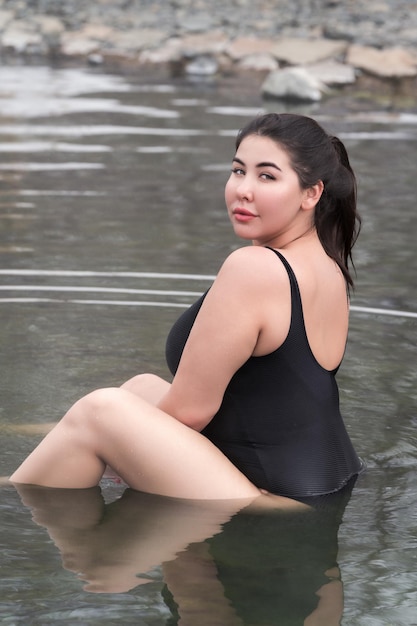 Übergewichtige junge Frau im schwarzen Badeanzug, die im Außenpool im Spa sitzt und über die Schulter schaut