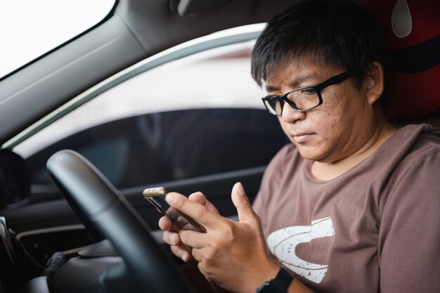 Übergewichtiger asiatischer Mann mit Brille ohne Sicherheitsgurt, der auf das Smartphone schaut, um die Textnachricht während des Staus zu überprüfen, gefährliches Fahren, Ablenkung von der Aufmerksamkeit kann einen Autounfall verursachen