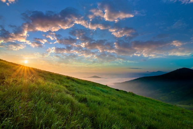 Berglandschaft bei schönem Wetter bei Sonnenaufgang. Grüner grasartiger steiler Hügel, nebeliges Tal und entfernte Berge unter hellem blauem Himmel mit beleuchtet, indem man weiße Wolken der Sonne anhebt. Schönheit des Naturkonzeptes.