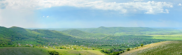 Berglandschaft, Wiese mit grünem Rasen, kleines Dorf und Hügel in der Ferne, weiße Wolken im Himmel